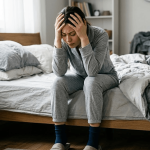 A person sitting on the edge of a bed holding their head to stabilize themselves after experiencing a dizzy spell